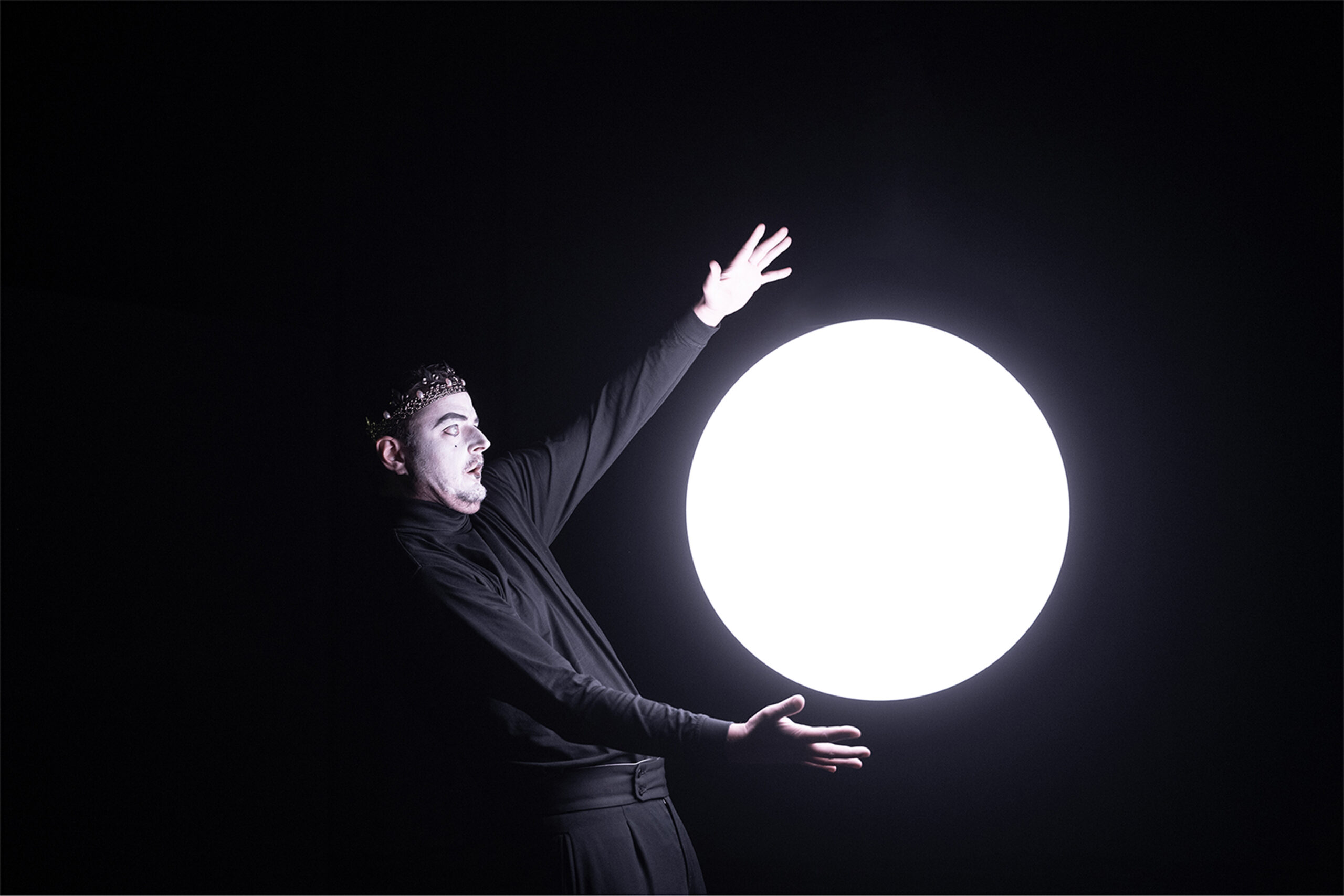 Ein schwarz gekleideter Mann mit weiß bemaltem Gesicht und einer Krone auf dem Kopf rahmt mit den Armen ein großes rundes Licht ein, das wie der Vollmond anmutet.