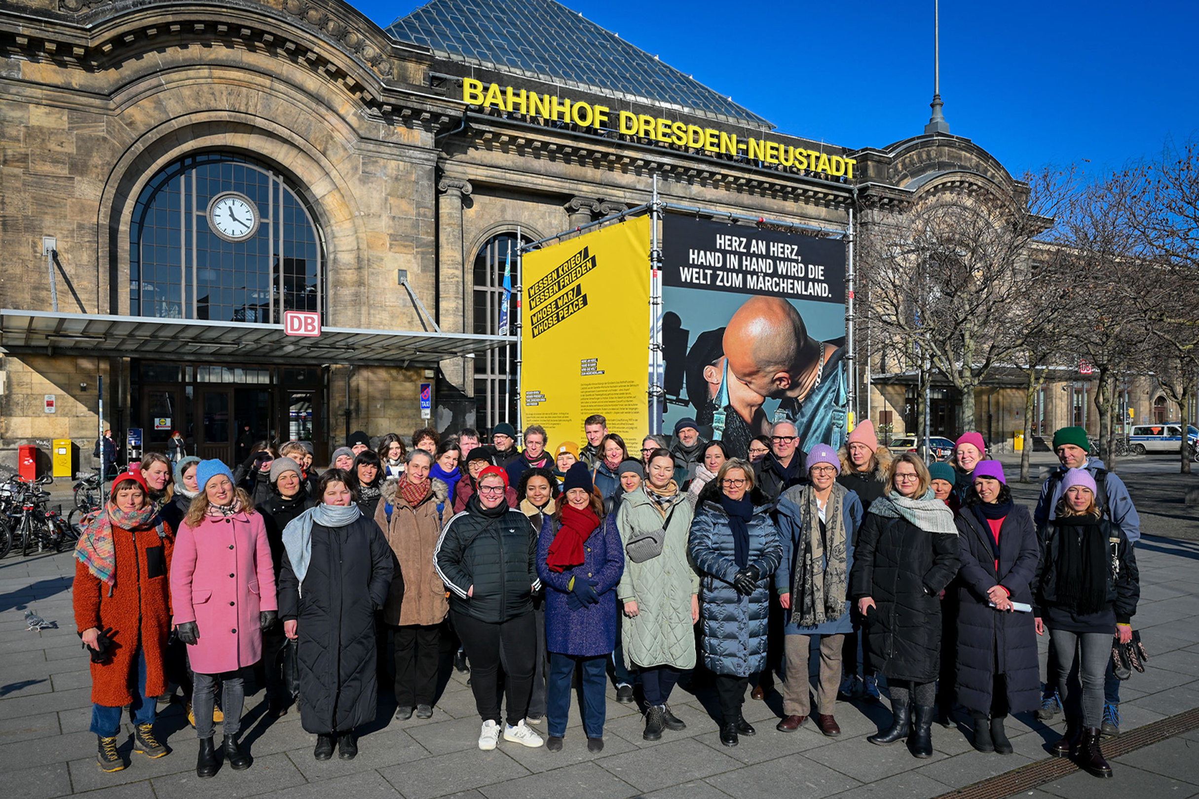 Website_WOD_(c)Anja-Schneider Eine große Personengruppe steht vor dem Bahnhofsgebäude Dresden-Neustadt. Auf dem Platz ist auch eine Plakataktion zum Thema "Weltoffenes Dresden" zu sehen.