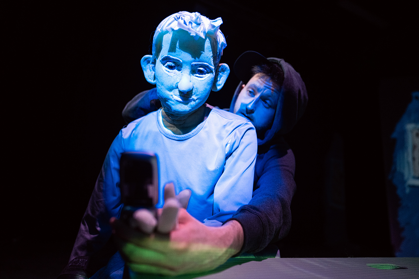 Ein Puppenspieler auf einer dunklen Bühne spielt eine lebensgroße Puppe eines Jugendlichen, der in blauem Licht sitzt und auf sein Smartphone schaut.