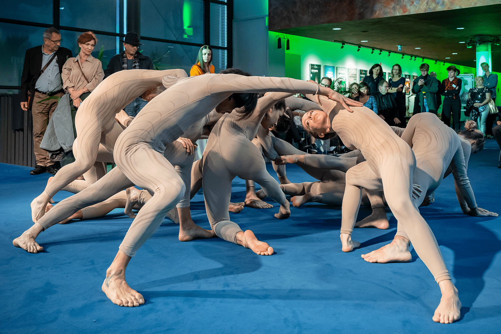 Tänzer in beigen Ganzkörperanzügen tanzen umgeben von Zuschauern eine Choreografie im Foyer.