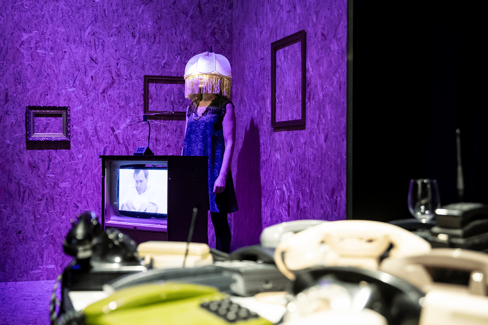 Eine Frau mit einem Lampenschirm auf dem Kopf steht in der Ecke eines Raumes. Ein laufender Fernseher und viele Telefone sind ebenfalls zu sehen.